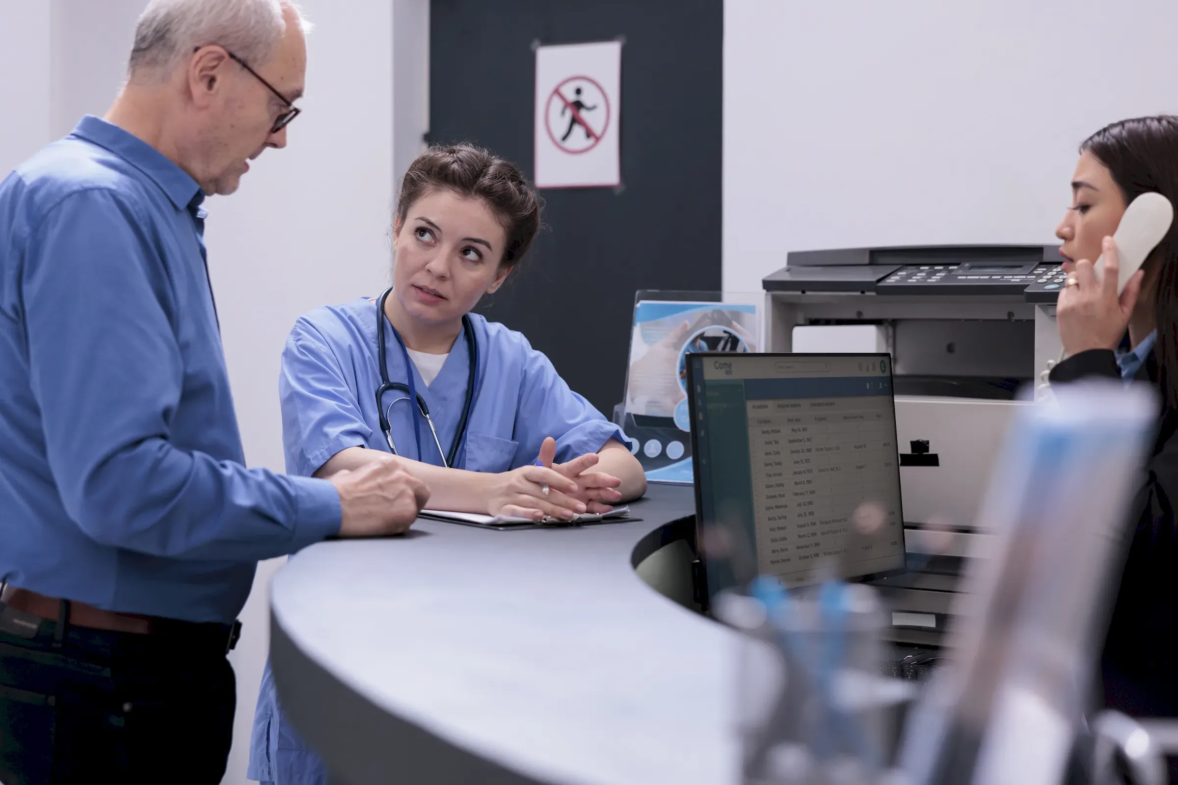 A clinician at a desk going through paperwork with a man, a colleague on the phone behind.