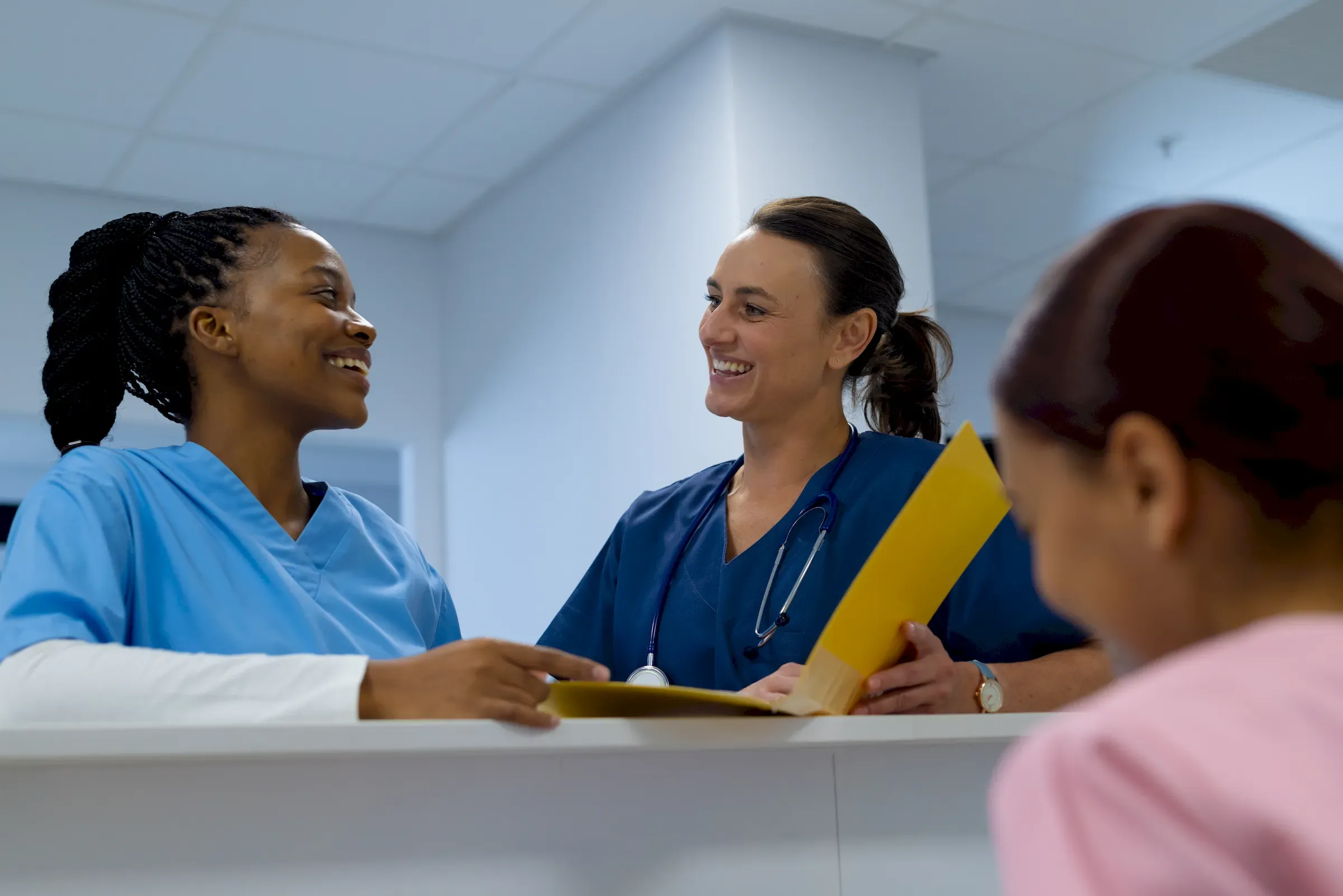 Two nurses at a hospital reception desk talking with someone across the counter.