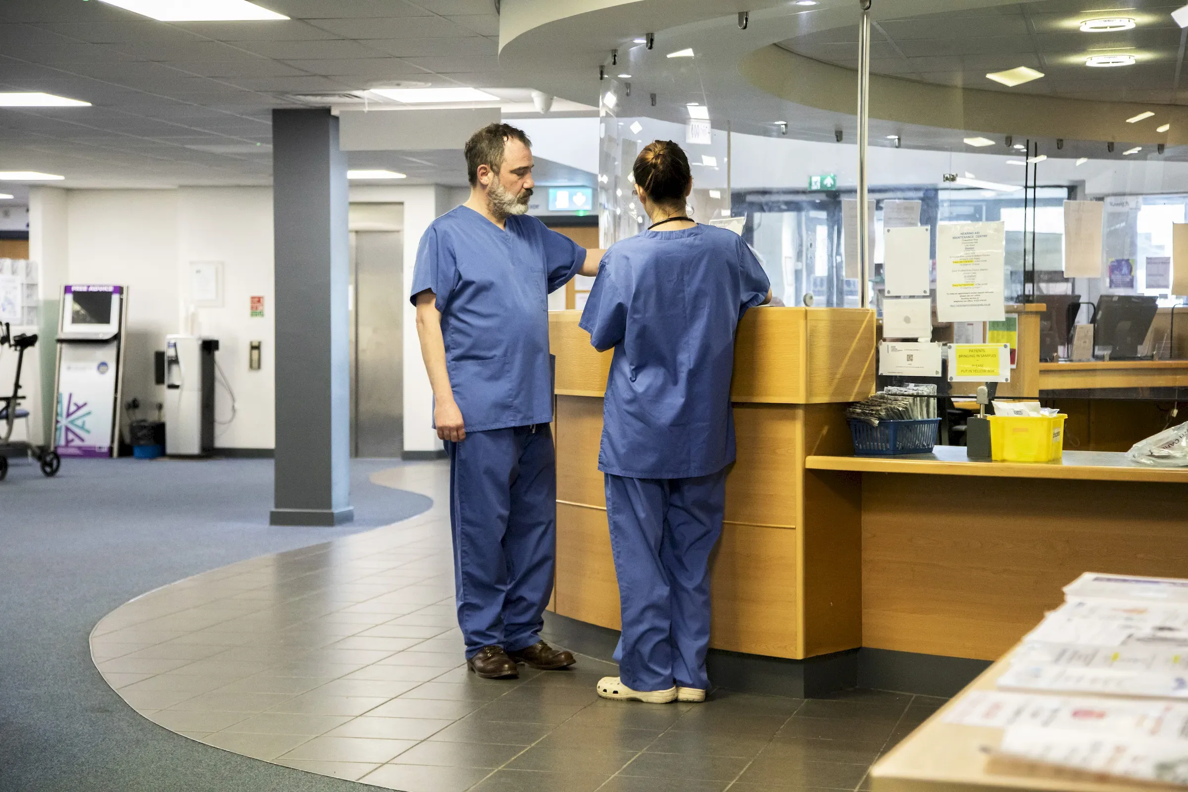 Two staff in scrubs at a hospital reception counter, one checking in at the desk.