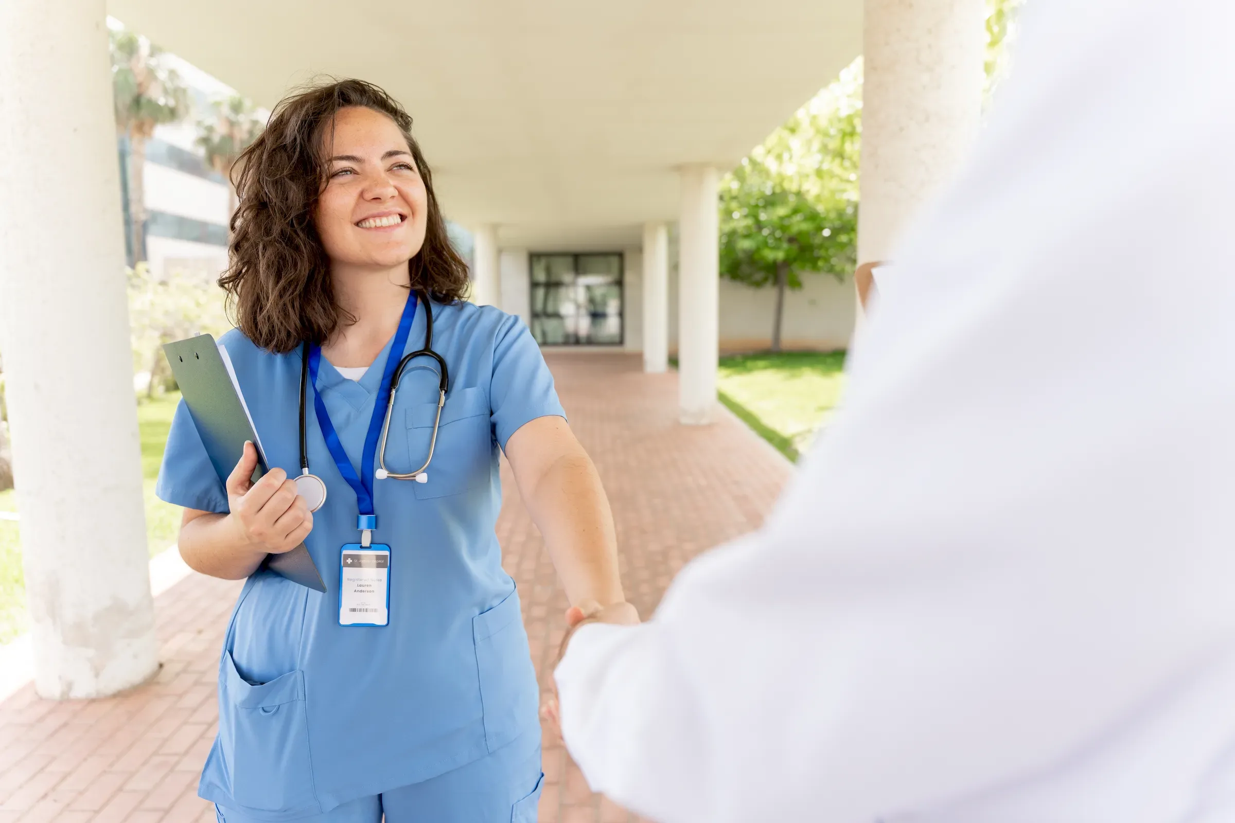 A clinician with a lanyard and tablet shaking hands with a colleague in a white coat.