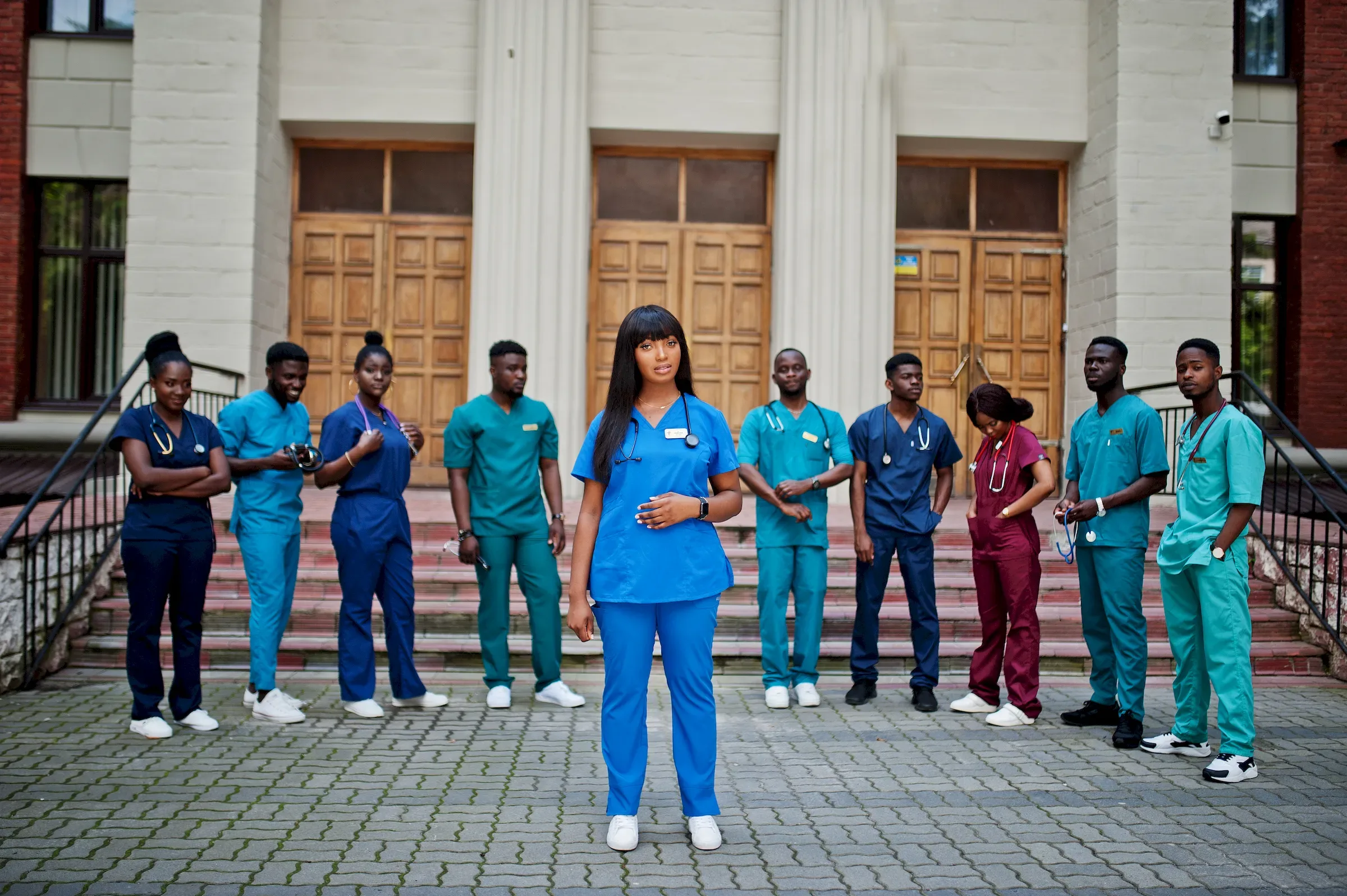 A group of clinical staff in coloured scrubs standing on the steps of a building, one in front.